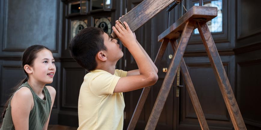 A brother and sister look through a wooden telescope indoors