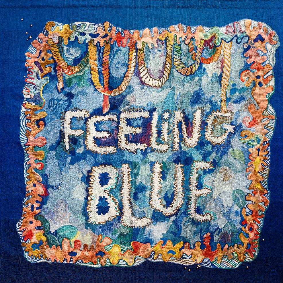 The artwork Feeling Blue. A brightly coloured tapestry featuring a blue border and the words 'Feeling Blue' in the centre. Shapes resembling coral, rope and waves frame the central design