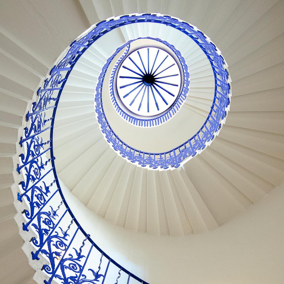 A photo looking up through a spiral staircase with ornate blue metal railings. The stairs appear to swirl up into a point in the ceiling