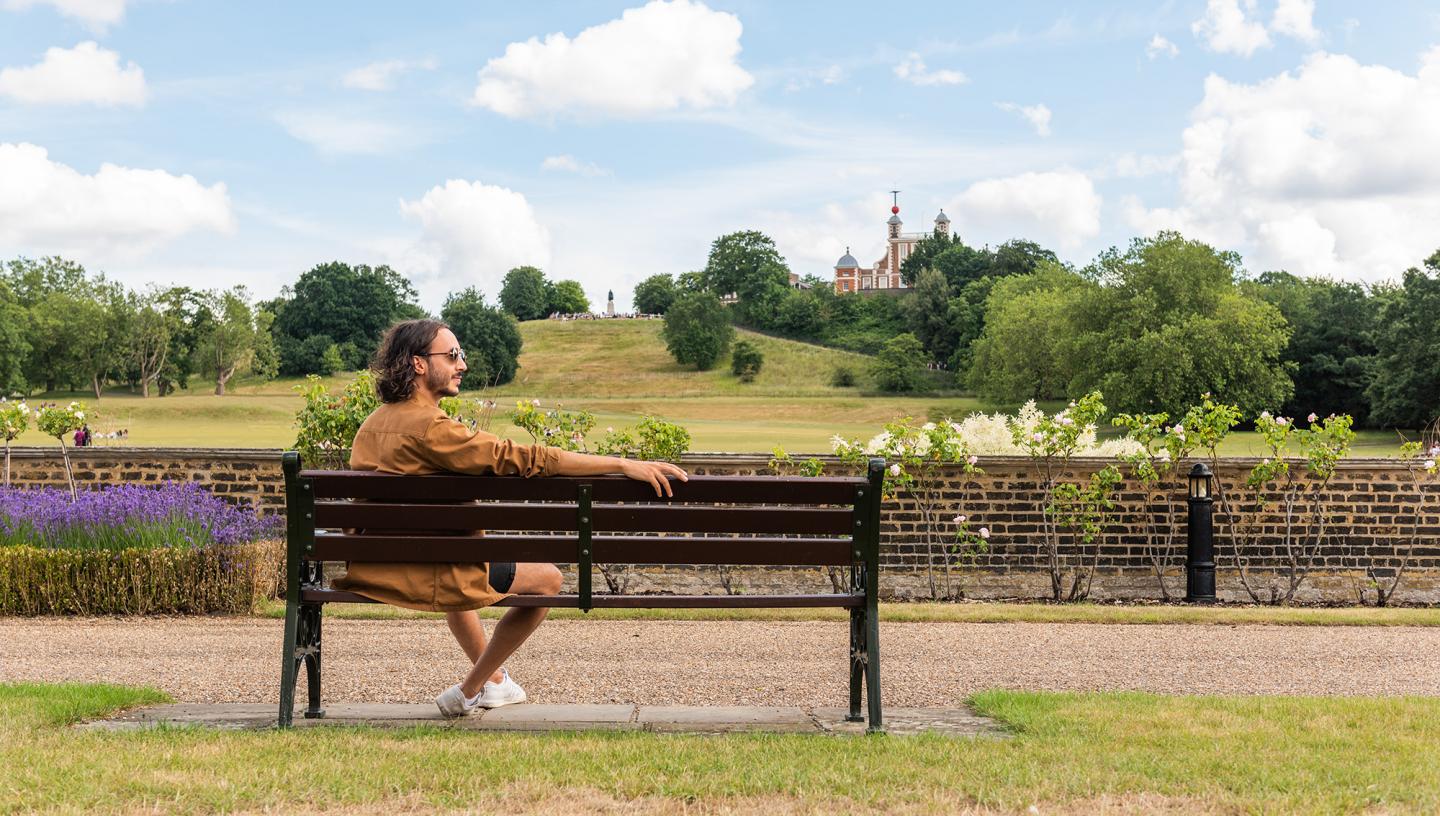 A man sits on a park bench beside Greenwich Park, with the Royal Observatory in the background