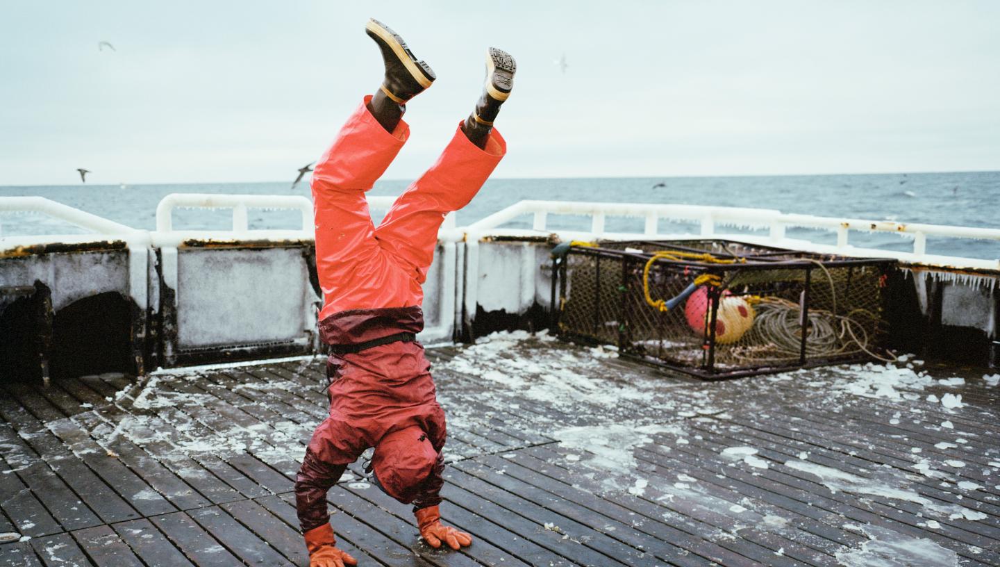 Image shows a man in an orange work suit doing a handstand on an icy deck