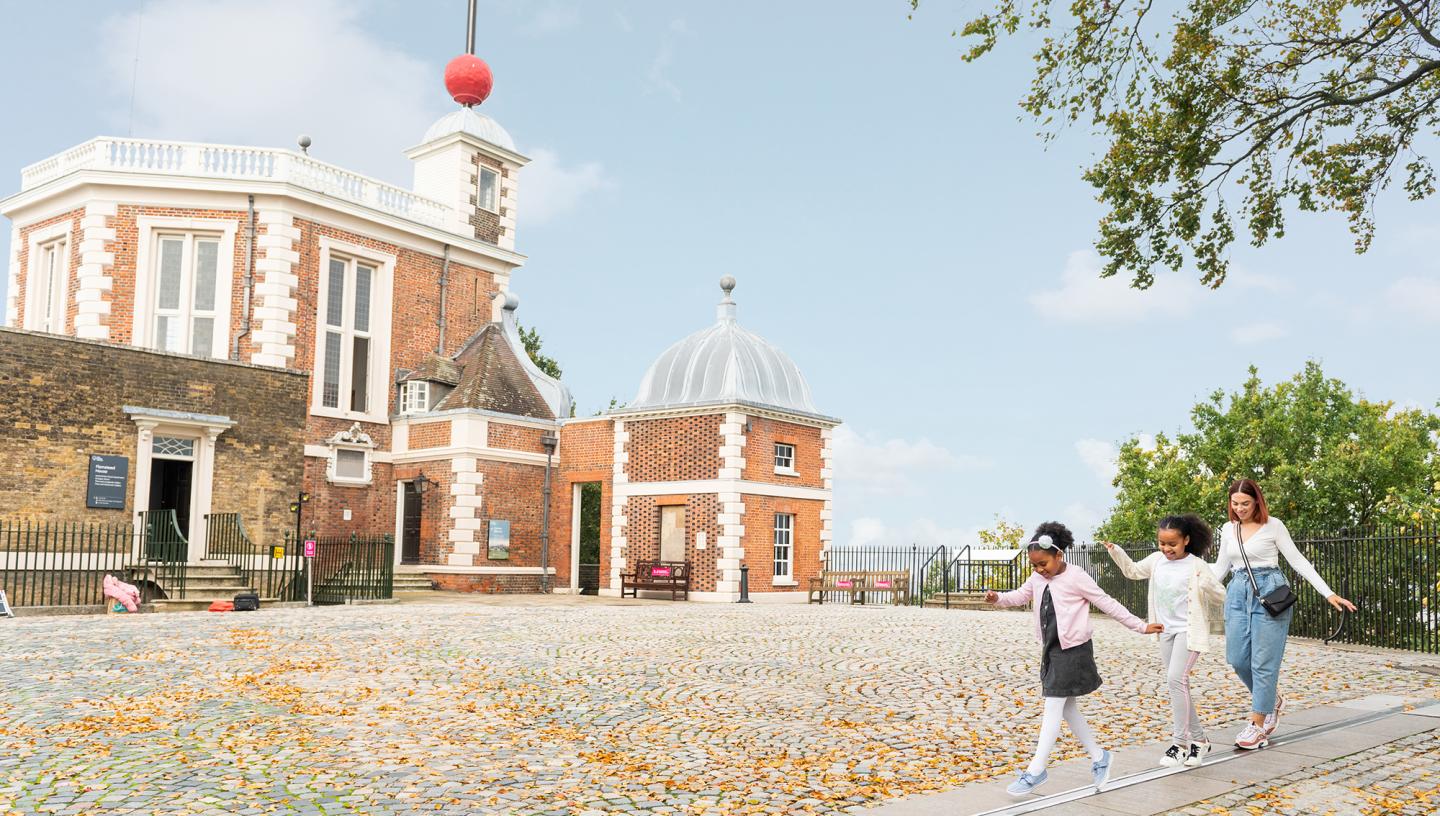 A family walk along the Prime Meridian line at Greenwich, with the historic Royal Observatory buildings in the background