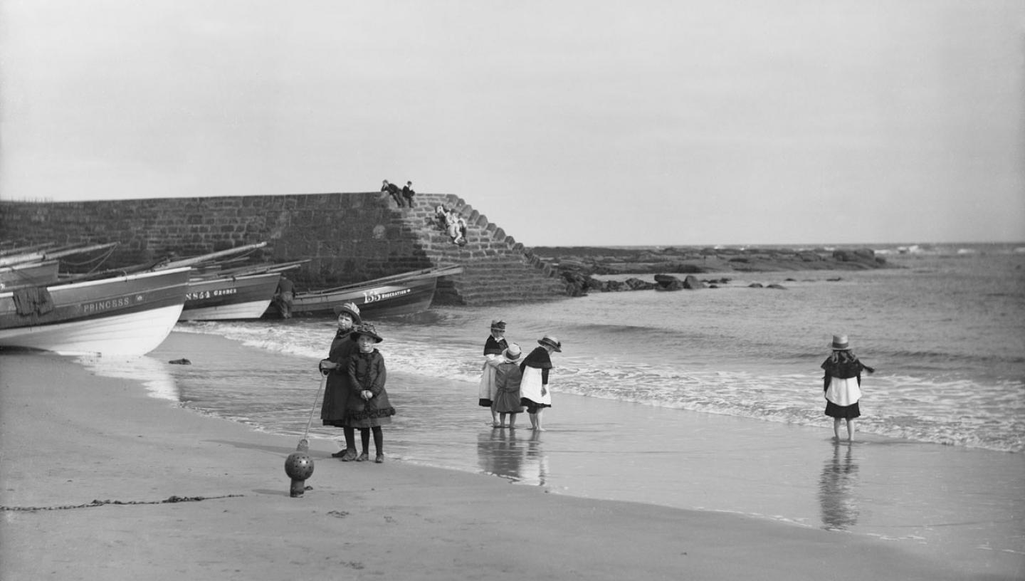 A black and white historic photograph of a family on the beach, with fishing boats and a harbour wall in the background
