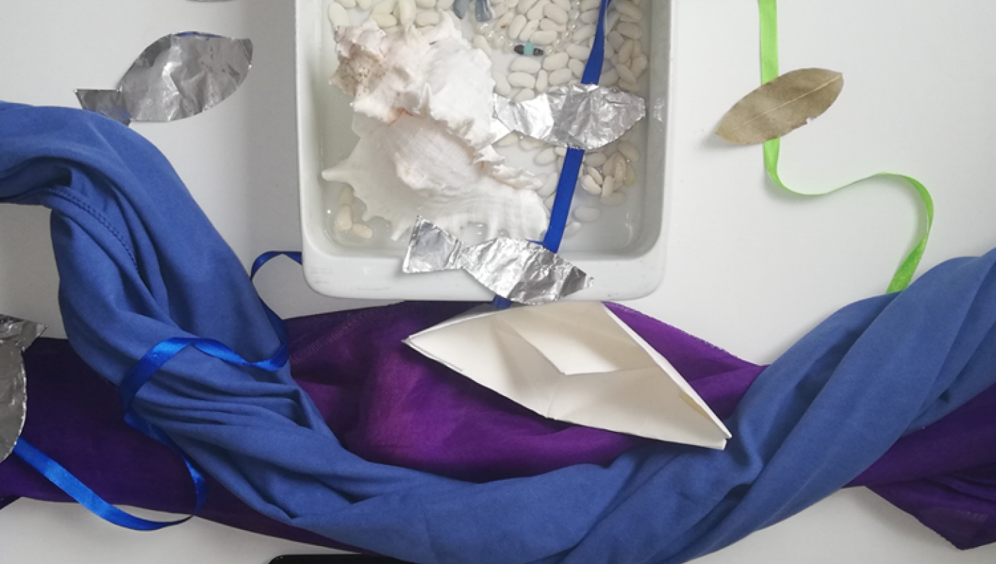 Final sensory tray showing tray filled with shells, dried beans, leaves, ribbons, foil fish on either side, blue and purple coloured fabric stretched out like a river with a paper boat and a tablet playing a video