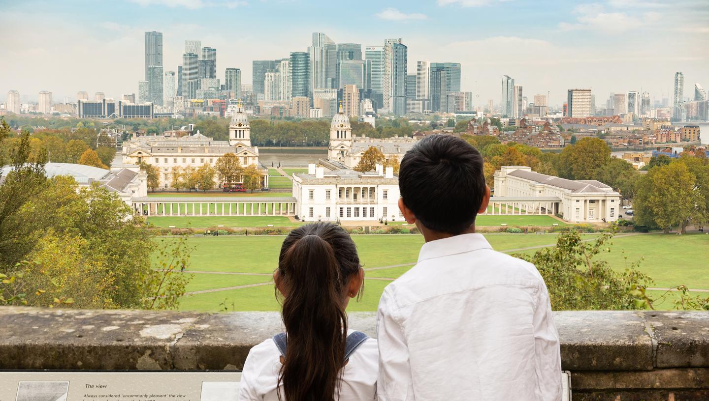 A brother and sister with their back to the camera look out from the viewpoint at the top of Greenwich Park towards historic Greenwich