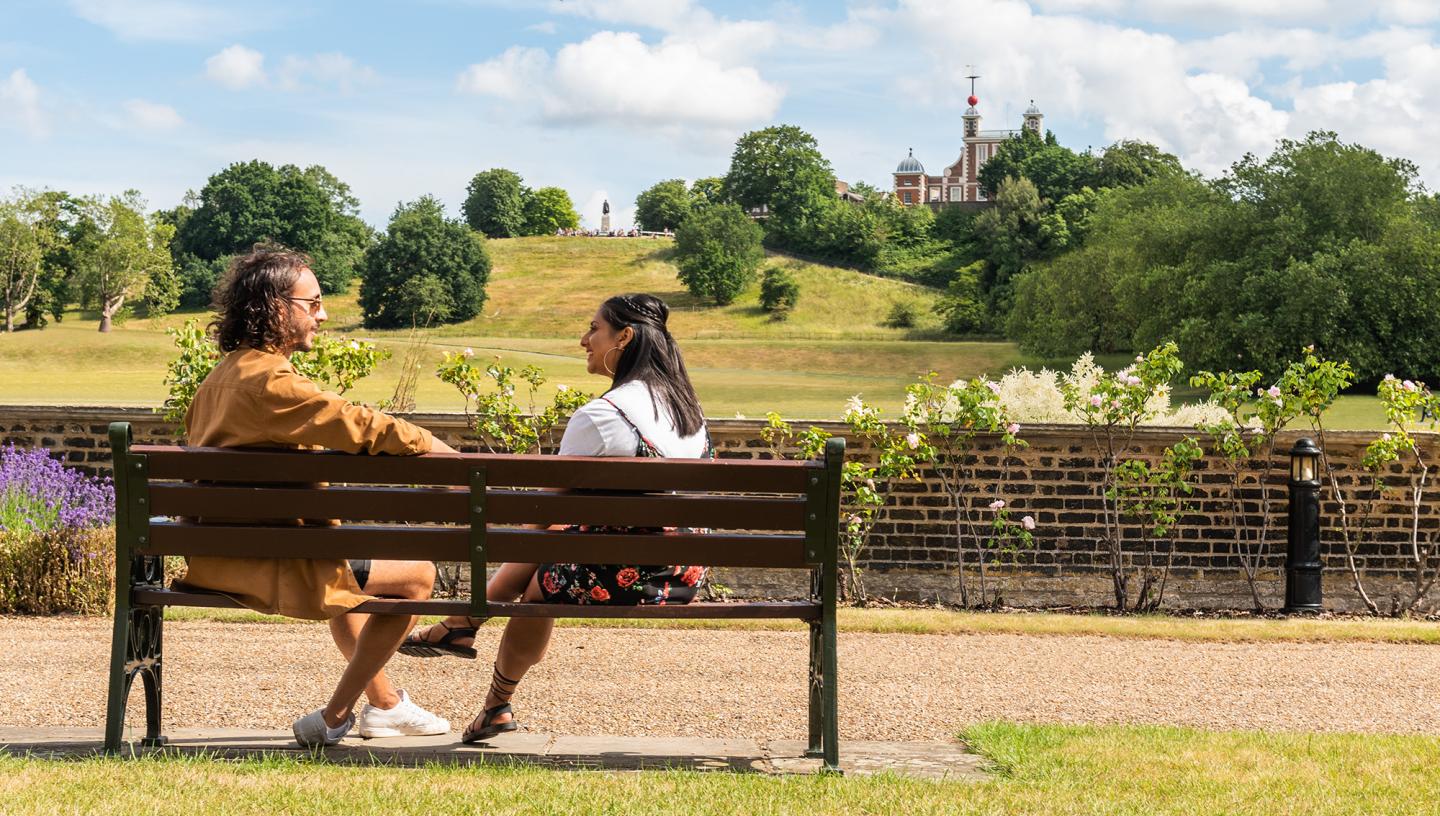 A couple sits on a bench in the sun, with Greenwich Park and the Royal Observatory in the distance