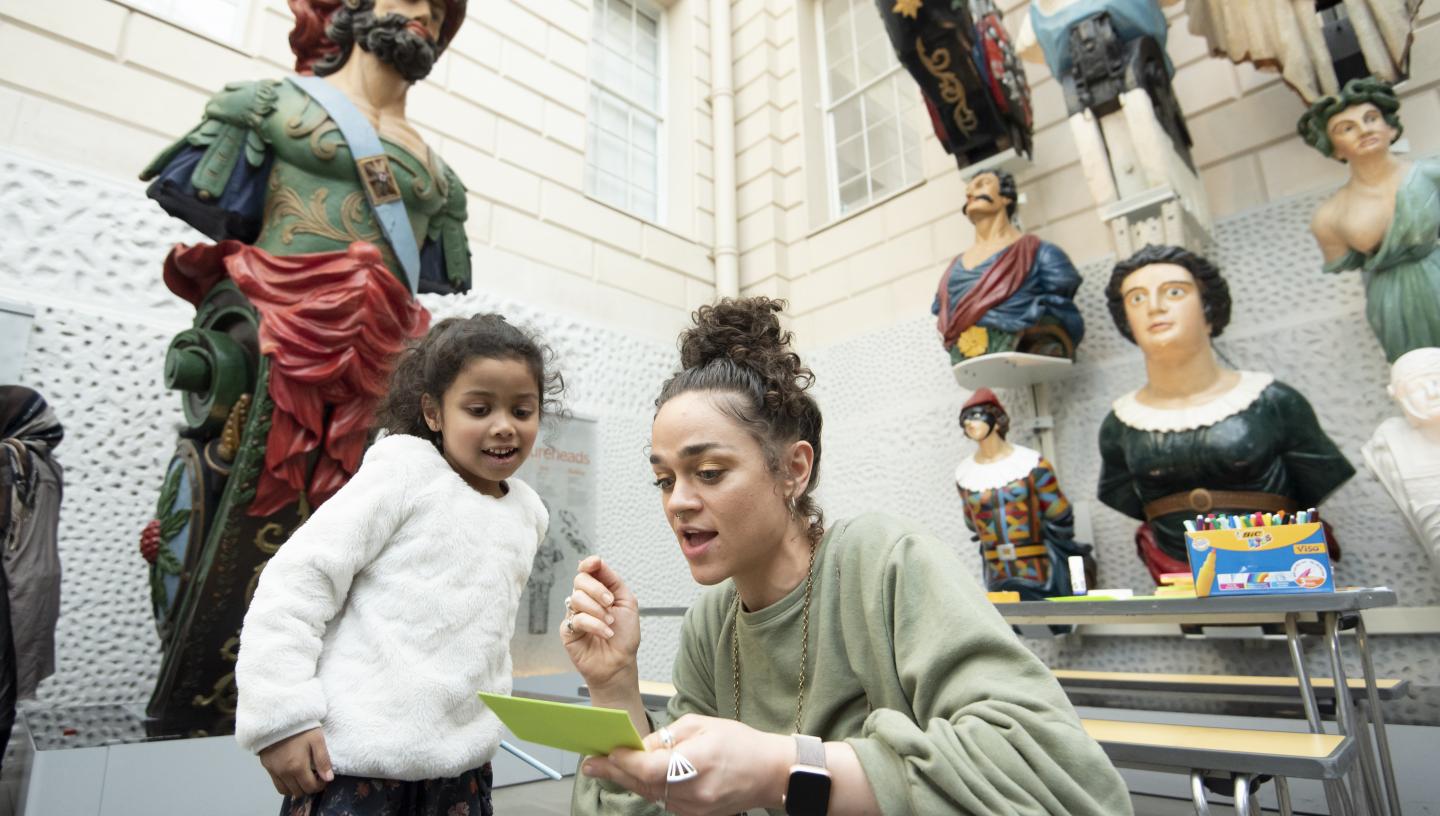 An adult and child are looking at a piece of paper. In the background are figureheads.