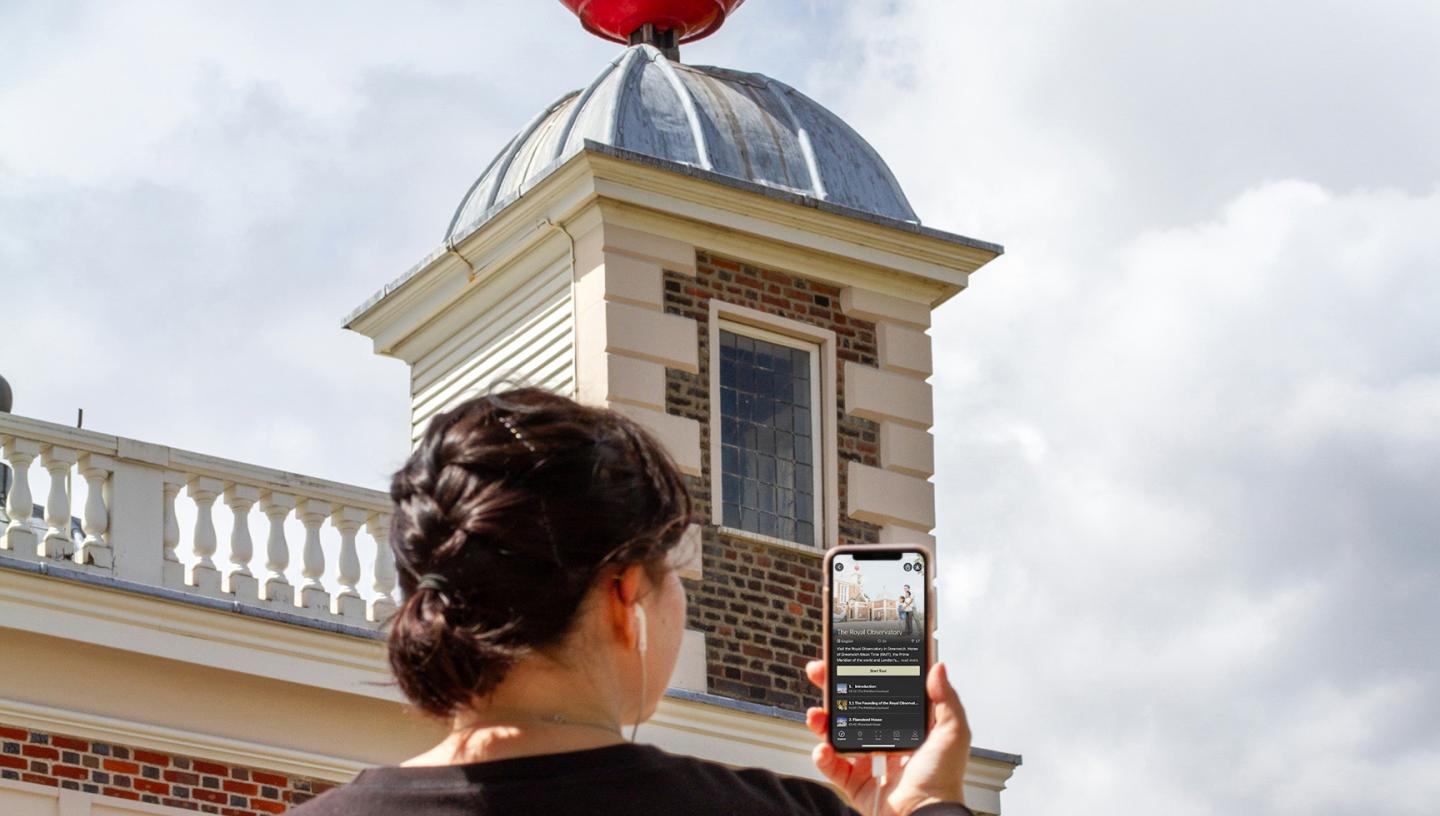 A woman holds her phone as she listens to the Royal Observatory audio tour, with the Observatory buildings in the background