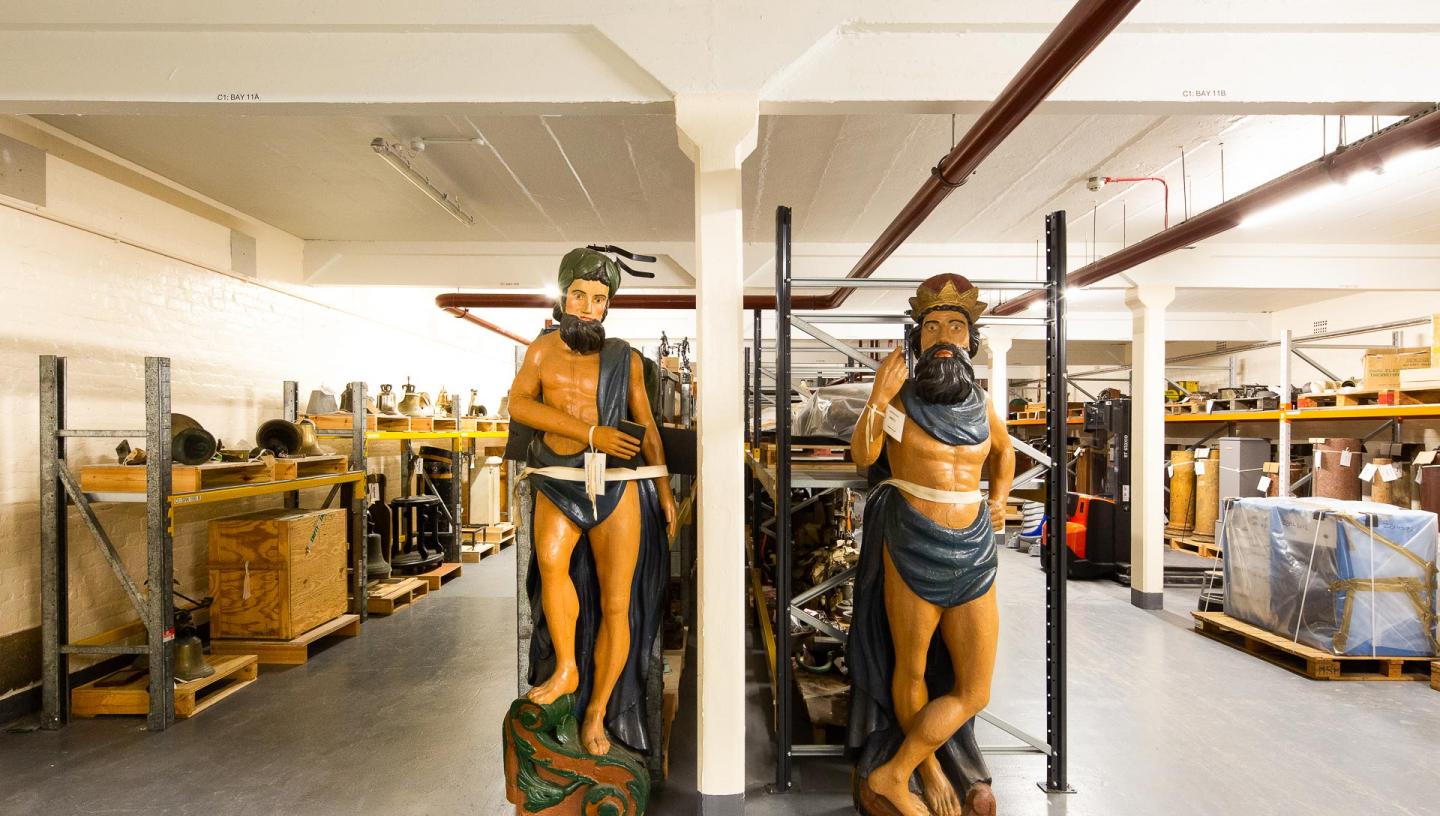 Inside Royal Museums Greenwich collections centre - rows of objects including two ships figureheads
