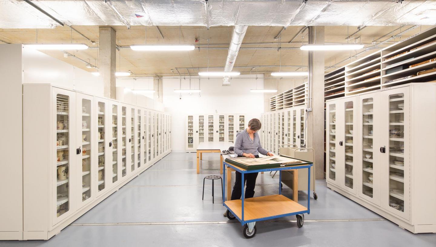 A woman stands at a desk in the middle of a museum storage centre. She is surrounded by white storage and display cases