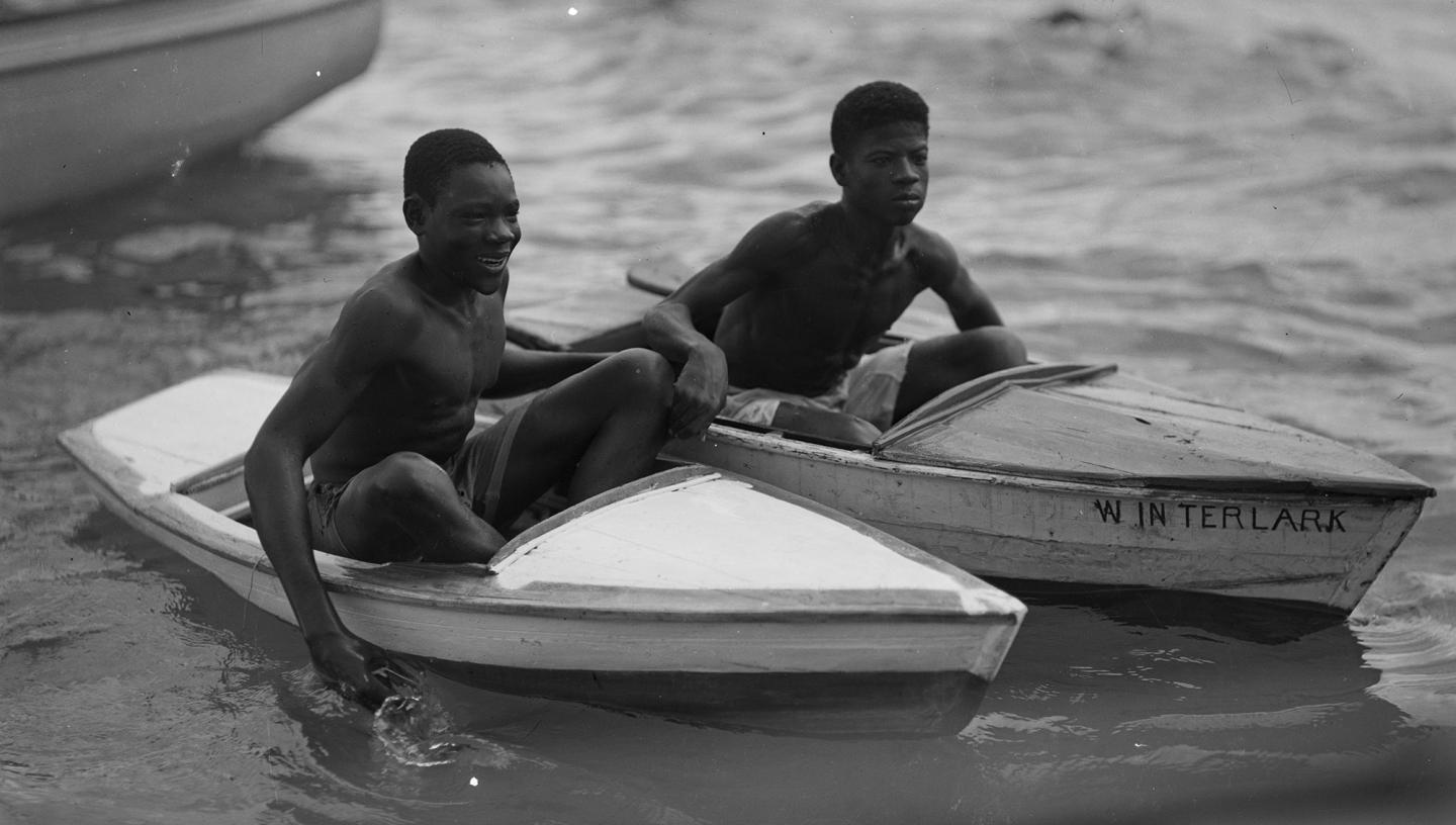 Historic black and white photo of boys in boats in Castries harbour, St. Lucia, West Indies. The negative is captioned 'Boys will be boys'