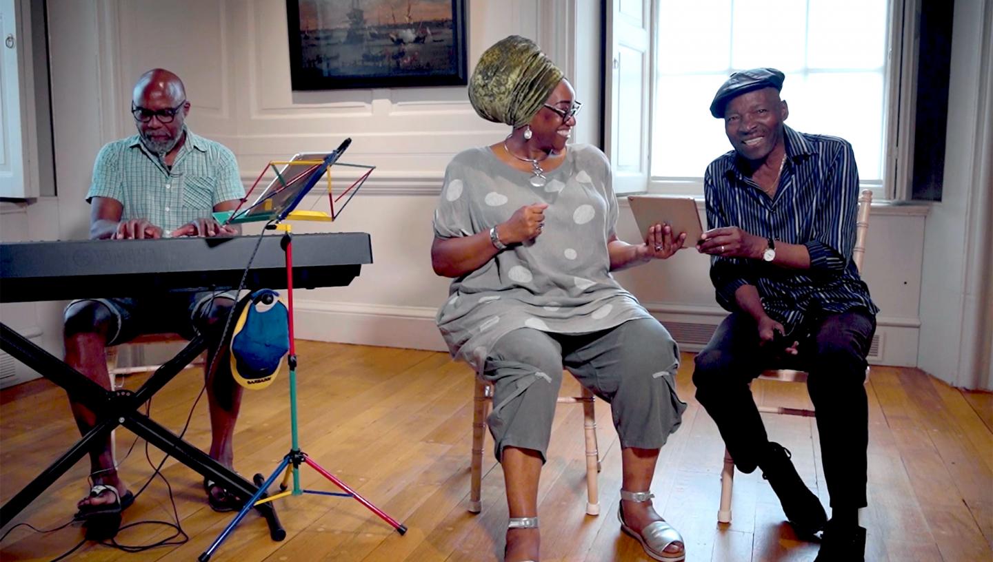 A group singing together, with a man playing a keyboard on the left and a woman and man sharing an iPad screen and singing on the right