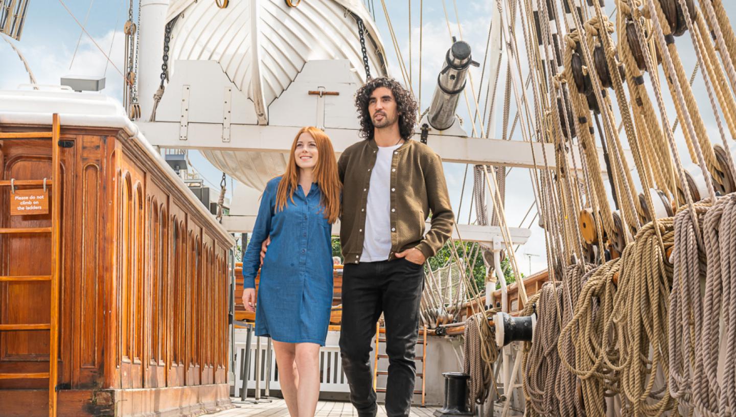 A couple walks along the main deck of historic ship Cutty Sark in summer, with rigging and masts above them