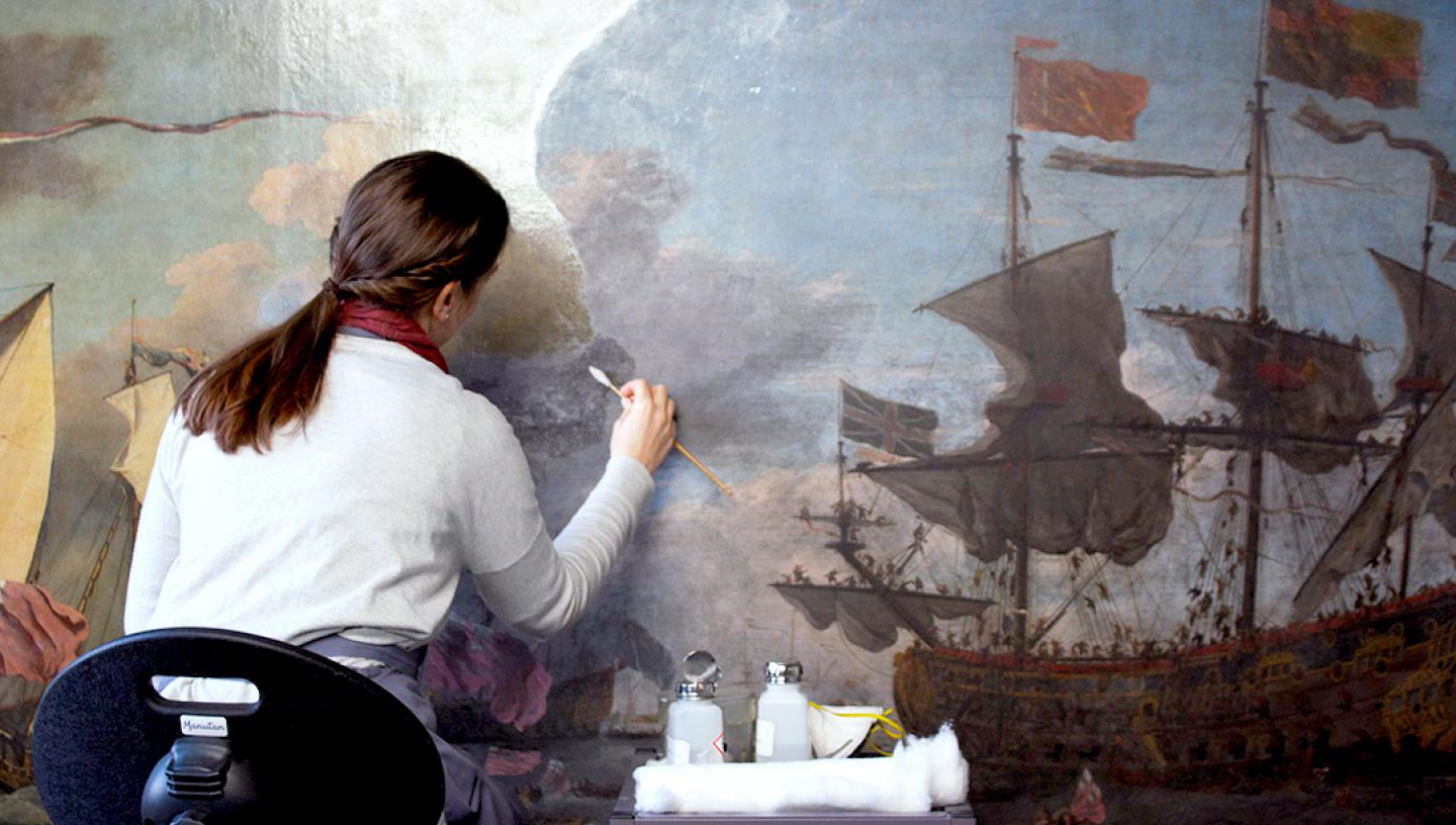 A conservator works on a small portion of a large maritime oil painting