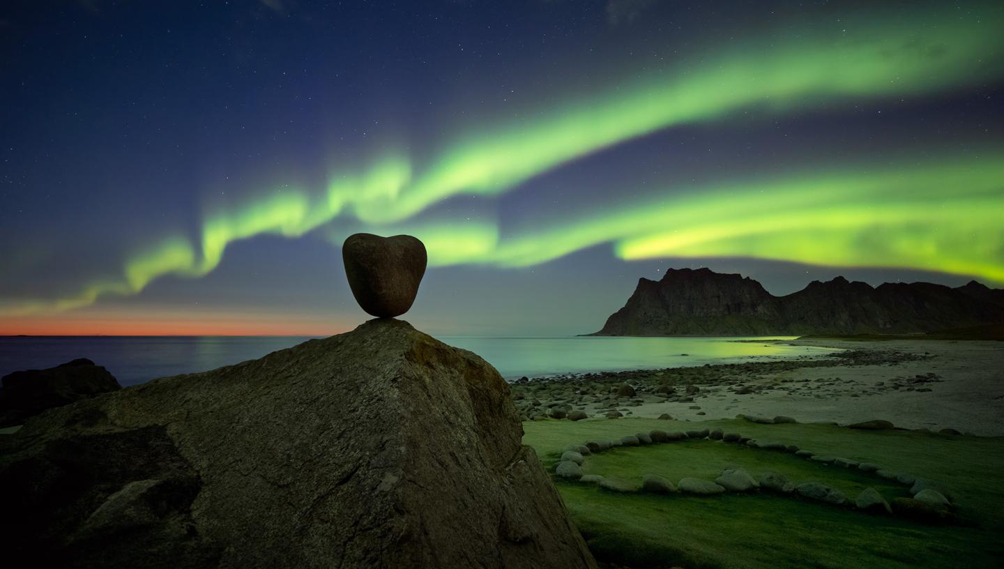 A stone in the shape of a heart is placed in front of a sky filled with green aurorae