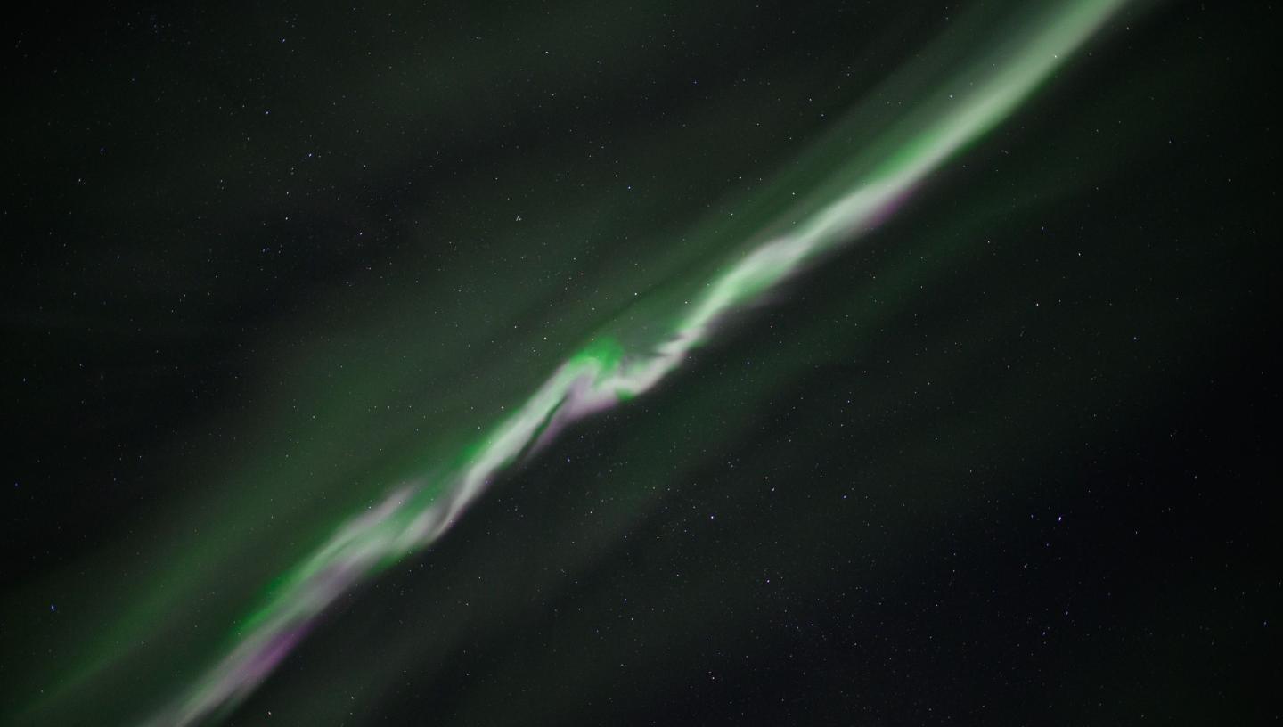 A thin blurred line of green and purple aurorae cuts diagonally across a dark sky
