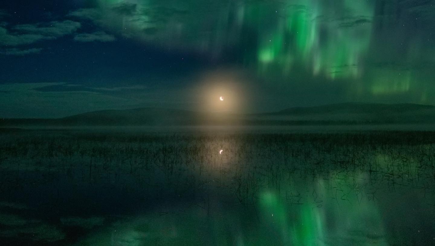 A green aurora reflected in water, with the Moon rising in the centre