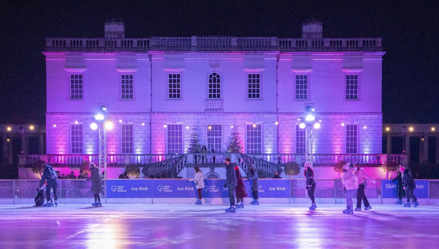 A photograph of the Queen's House Ice Rink at night, with the House and rink lit by purple lighting and skaters in the foreground