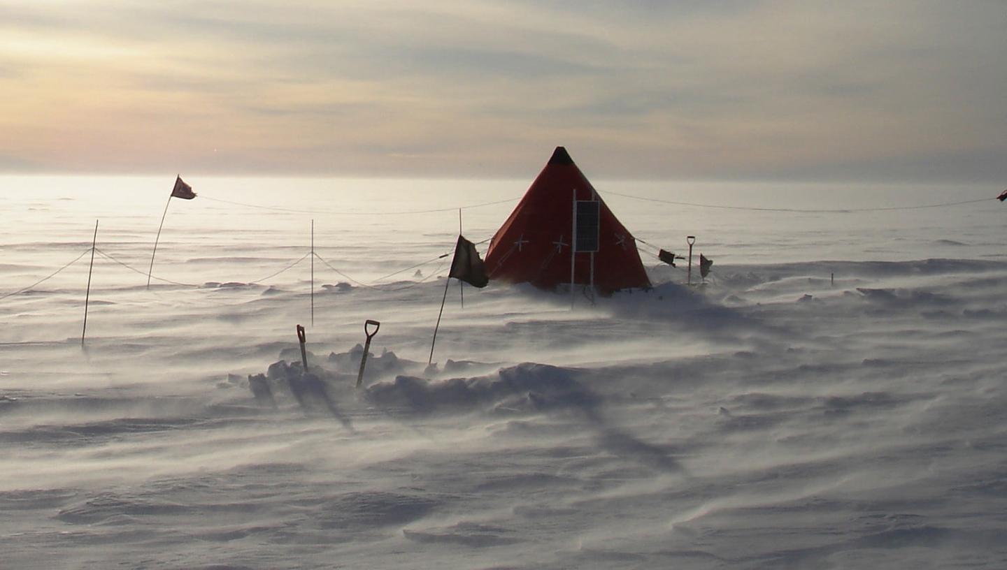 A pyramid tent in shadow pitched on a frozen glacier, with the wind picking up the snow on the ground