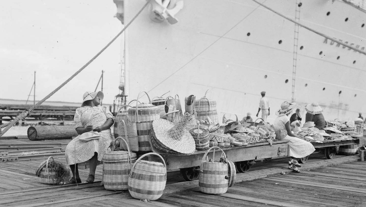 A black and white photo of market stall with a Jamaican woman selling baskets in front of a cruise liner in the background