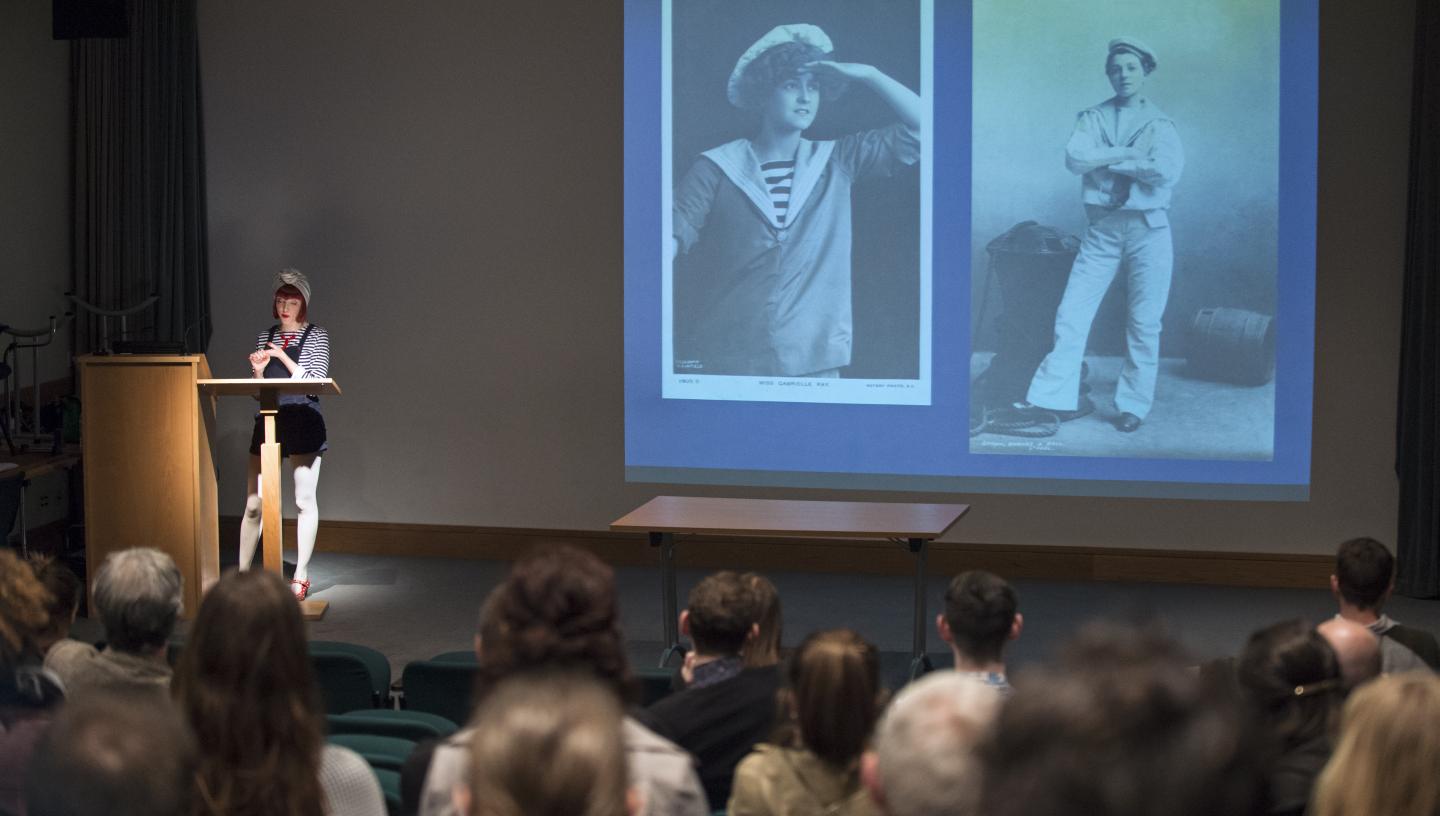 Photo of the Lecture Theatre at the National Maritime Museum filled with an audience watching a women present a talk on queer life at sea. Projected are two black and white photos of sailors, one wearing white bell bottoms and the other looking out to sea.