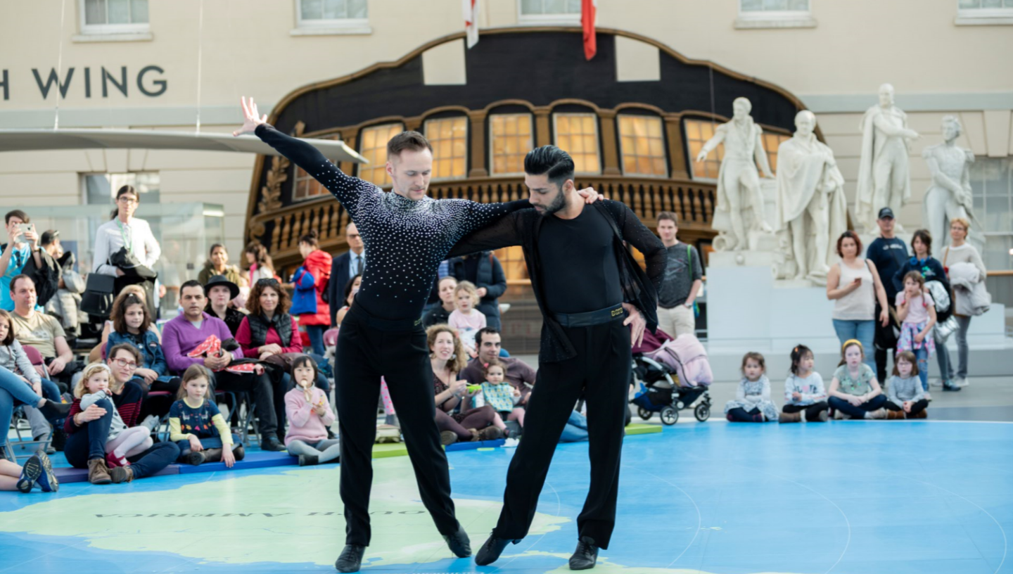 Photo of two male ballroom dancers  wearing black outfits, one with diamantes across his shoulders. A full audience sits behind the two dancers on the Great Map in the National Maritime Museum. 