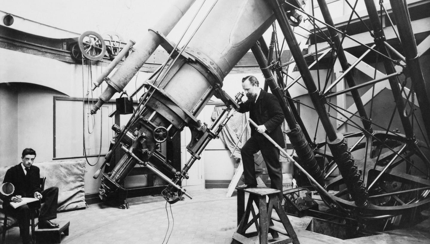 Historic photograph showing an astronomer peering into the eyepiece of the Great Equatorial Telescope at the Royal Observatory. Another astronomer is taking notes to the left