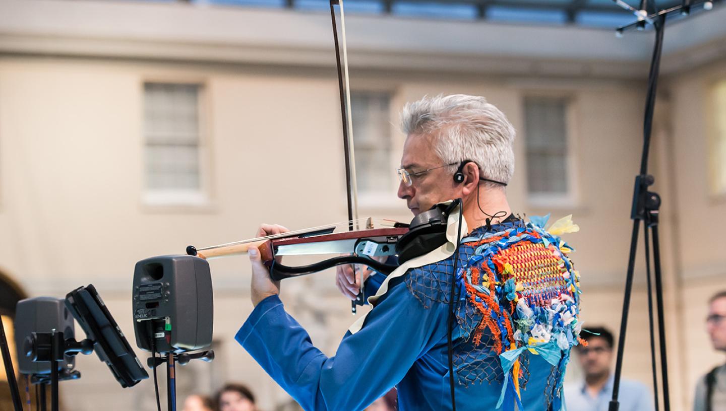A man plays a violin in the National Maritime Museum. He is wearing a blue denim jacket with decorative back panel made out of discarded ocean plastic