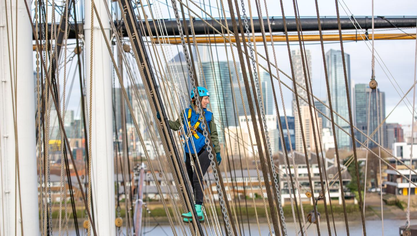 A woman climbing the rigging of historic sailing ship Cutty Sark, with the skyscrapers of Canary Wharf in the background