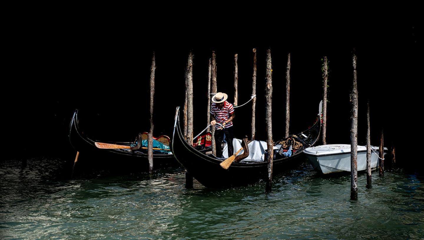 A gondolier ties up a gondola at a mooring poll in Venice. He is caught in the sunlight, but the rest of the photo is in shadow