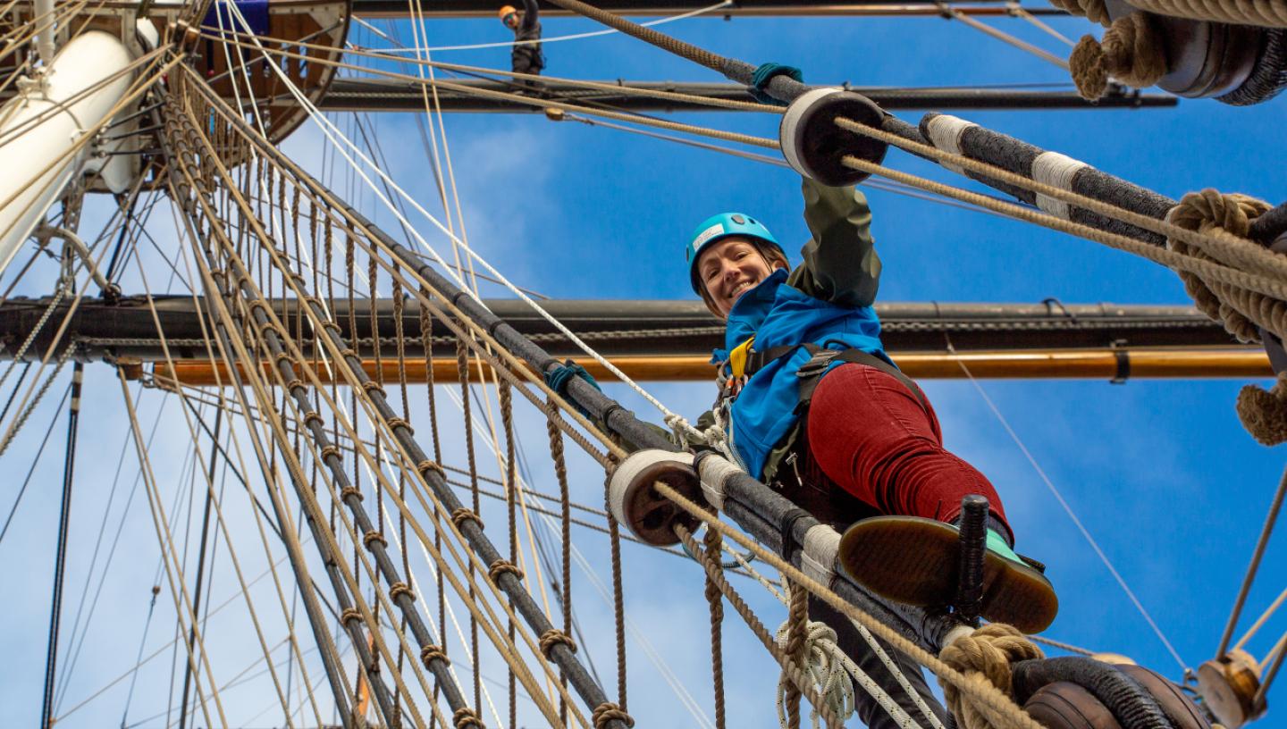 A woman climbs the rigging of historic sailing ship Cutty Sark. She is looking down at the photographer on deck, and the rigging and masts are above her