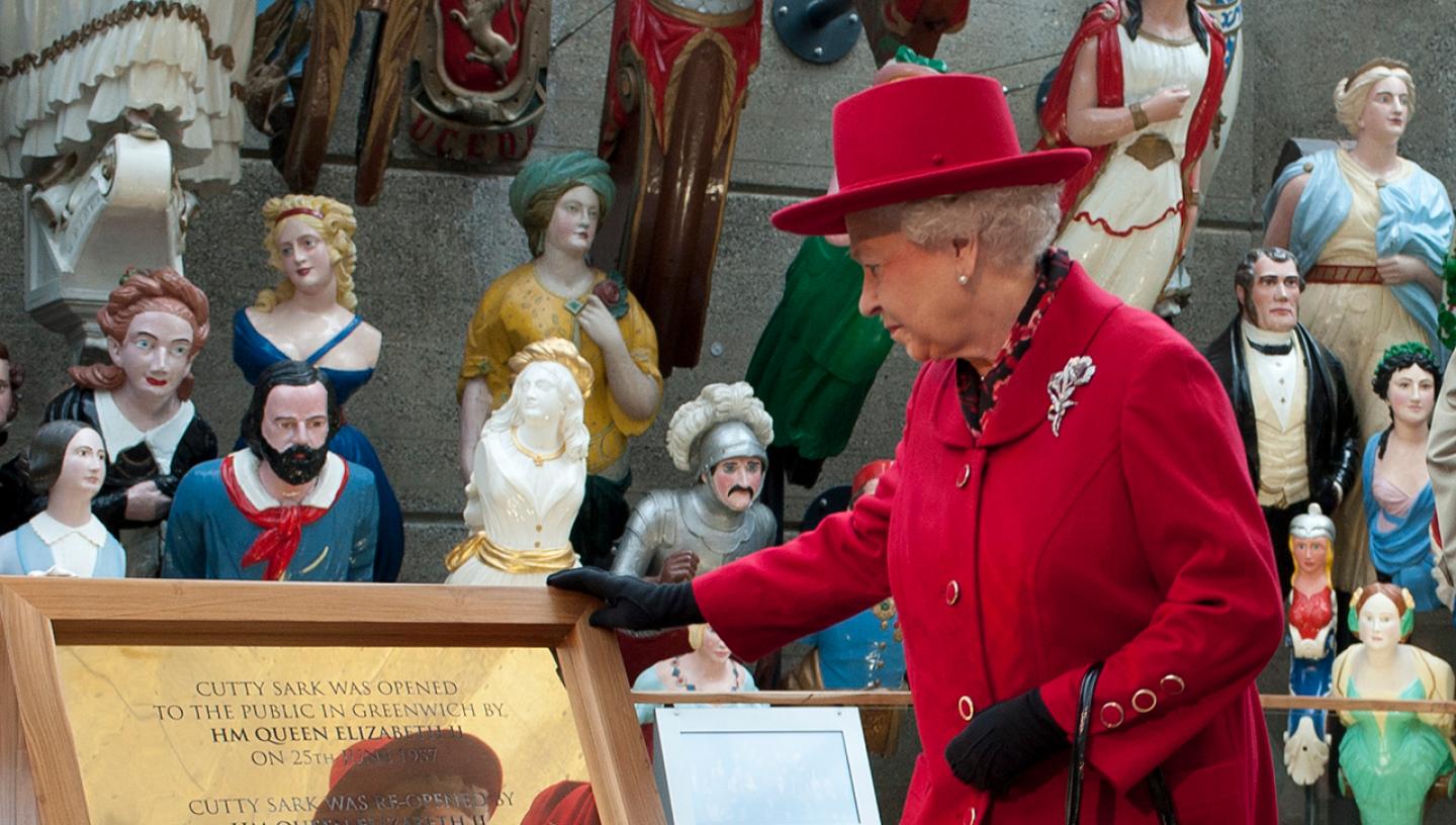 Her Majesty The Queen, wearing a red jacket and hat, examines a plaque dedicated to the opening of historic ship Cutty Sark
