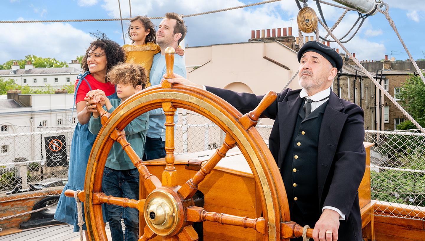 A smiling family of four look out on the main deck of Cutty Sark, with a character actor dressed as the ship's captain holding the ship's wheel