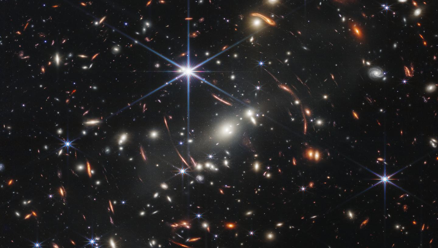 Thousands of small galaxies appear across this view. Their colors vary. Some are shades of orange, while others are white. Most appear as fuzzy ovals, but a few have distinct spiral arms. In front of the galaxies are several foreground stars. Most appear blue, and the bright stars have diffraction spikes, forming an eight-pointed star shape. There are also many thin, long, orange arcs that curve around the center of the image
