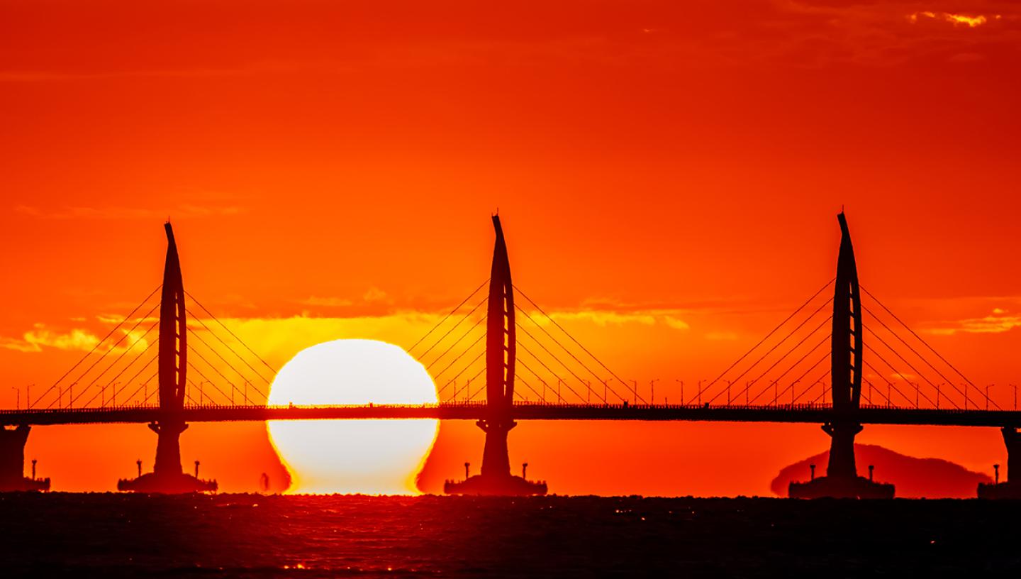 Image of a silhouette of a bridge in Hong Kong with orange and yellow sunset sky behind it, the Sun appears to be sitting on the water and the bottom of it is distorted in a mirage