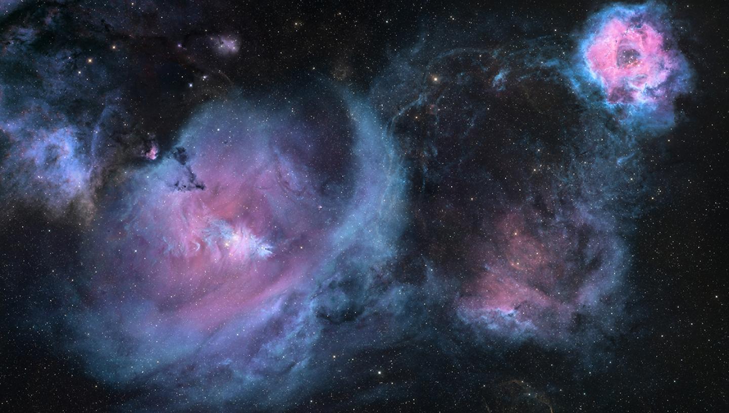 Rosette Nebula on right which resembles flower in blues, pinks and purples. Image shows dark black sky with stars and clouds of purple and light blue gases