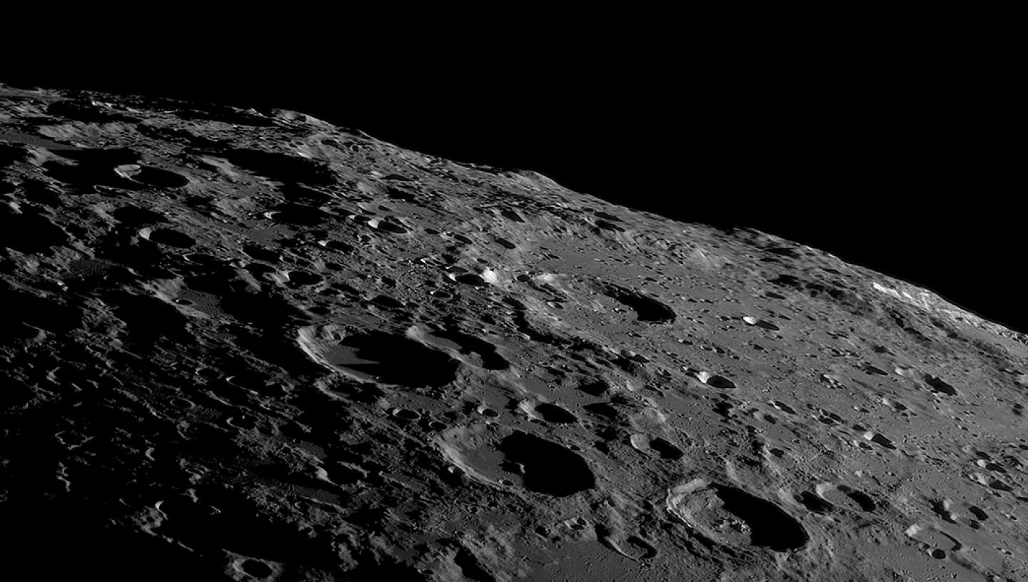 A black and white photograph of the Moon's surface, showing the rugged terrain in contrast against the black sky
