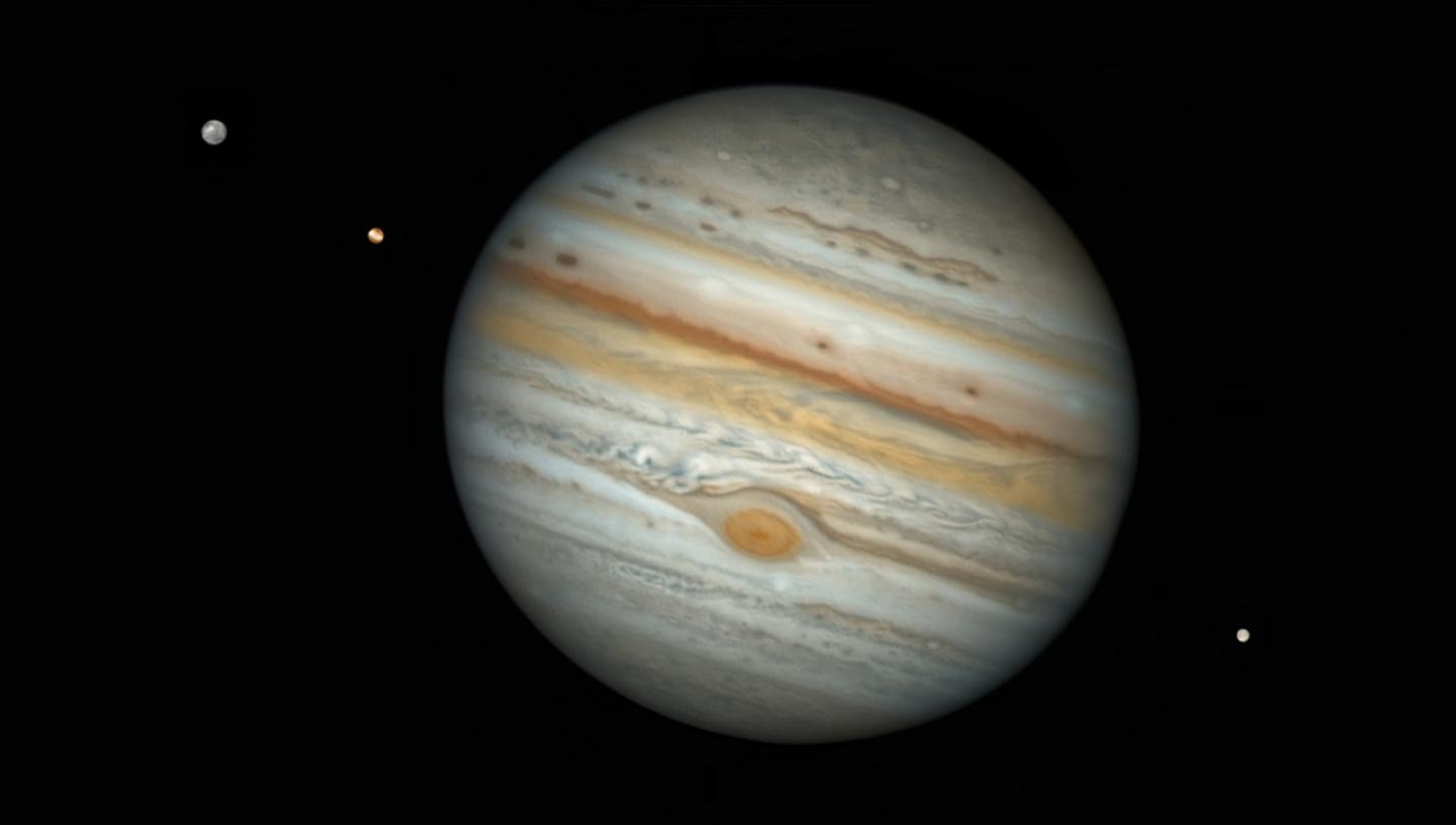 Image of Jupiter at a slight tilt, with three moons around it, two on a diagonal on the top left hand side and one on the bottom right hand side
