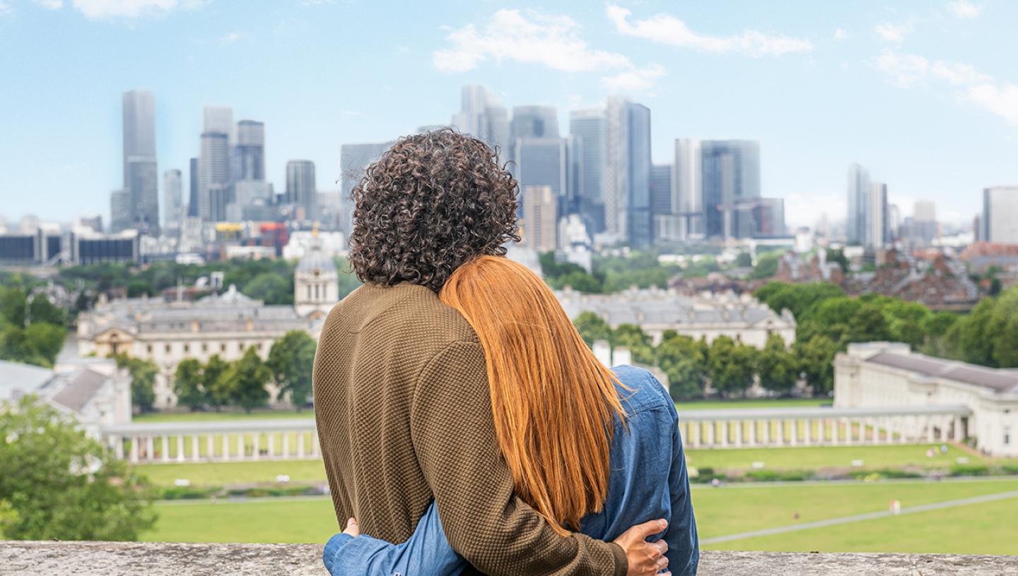 A man and a woman hug as they look out over historic Greenwich towards the skyscrapers of Canary Wharf. They have their backs to the camera, and the woman's head is nestled in the man's shoulder