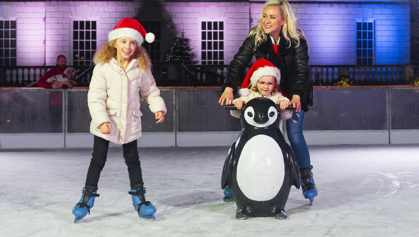 Image of ice rink at night time with house lit up in purple and blue lights behind three people, a mother with her two children, one is younger and using a penguin skate aid