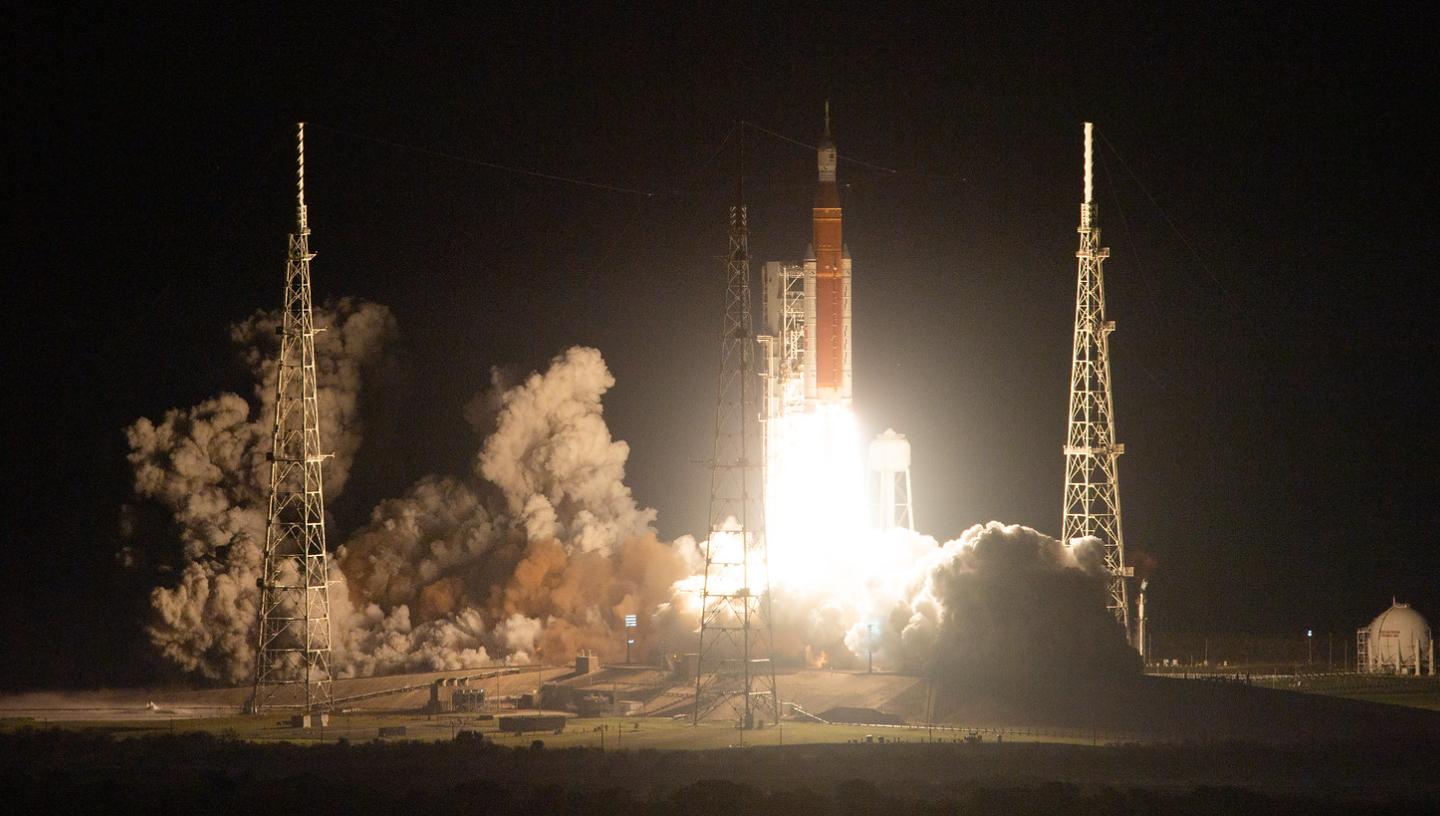 Image of orange rocket at night with white-hot flames coming out of the bottom and propelling it off the ground, a trail of grey smoke at the bottom