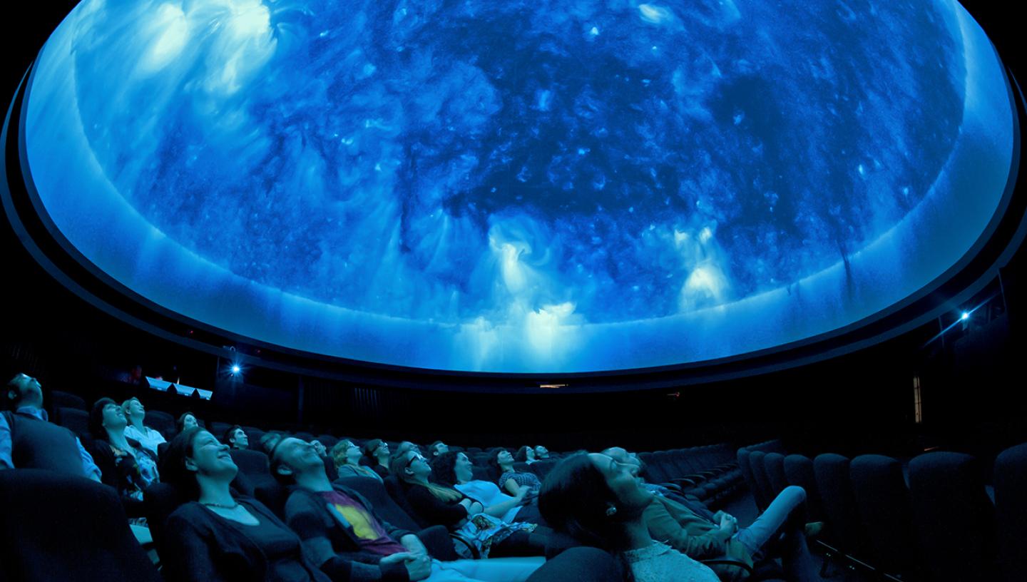 An audience enjoys a Planetarium show at the Royal Observatory. They are lying back in big armchairs, staring up at a ceiling filled with blue light projections of distant galaxies