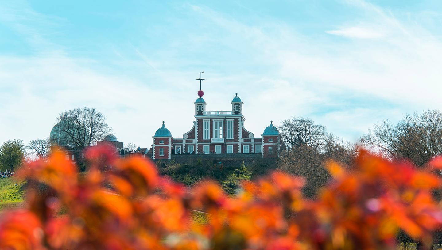 A photograph of the historic Royal Observatory Greenwich. The view is looking up at the brick building from Greenwich Park, with autumnal leaves in the foreground and a clear blue sky above