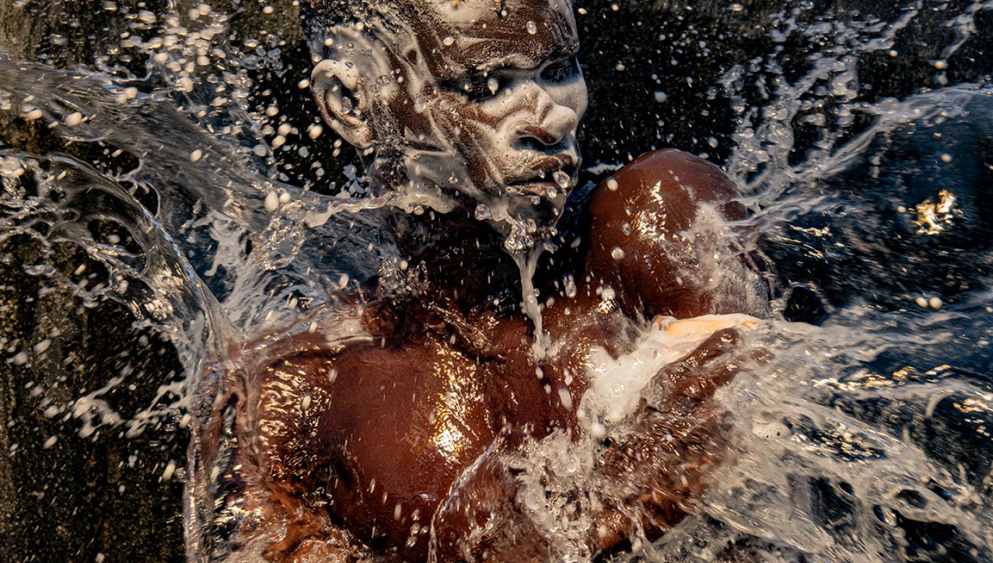 A photograph of a man washing. His face and hair is covered in soap suds, an water is being thrown over him