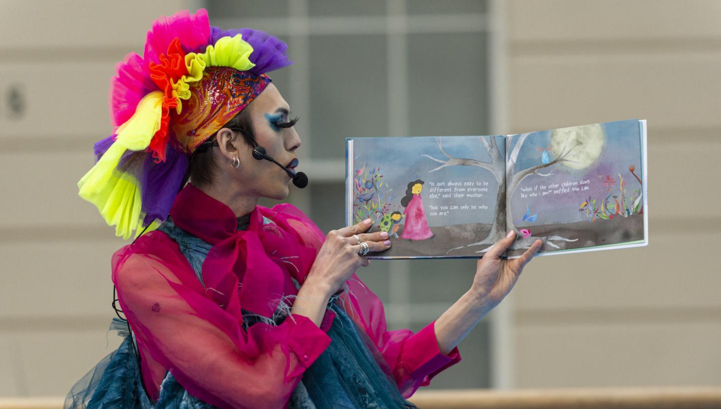 A Drag star with brightly coloured wig and nautical inspired dress reads from a picture book during a performance at the National Maritime Museum