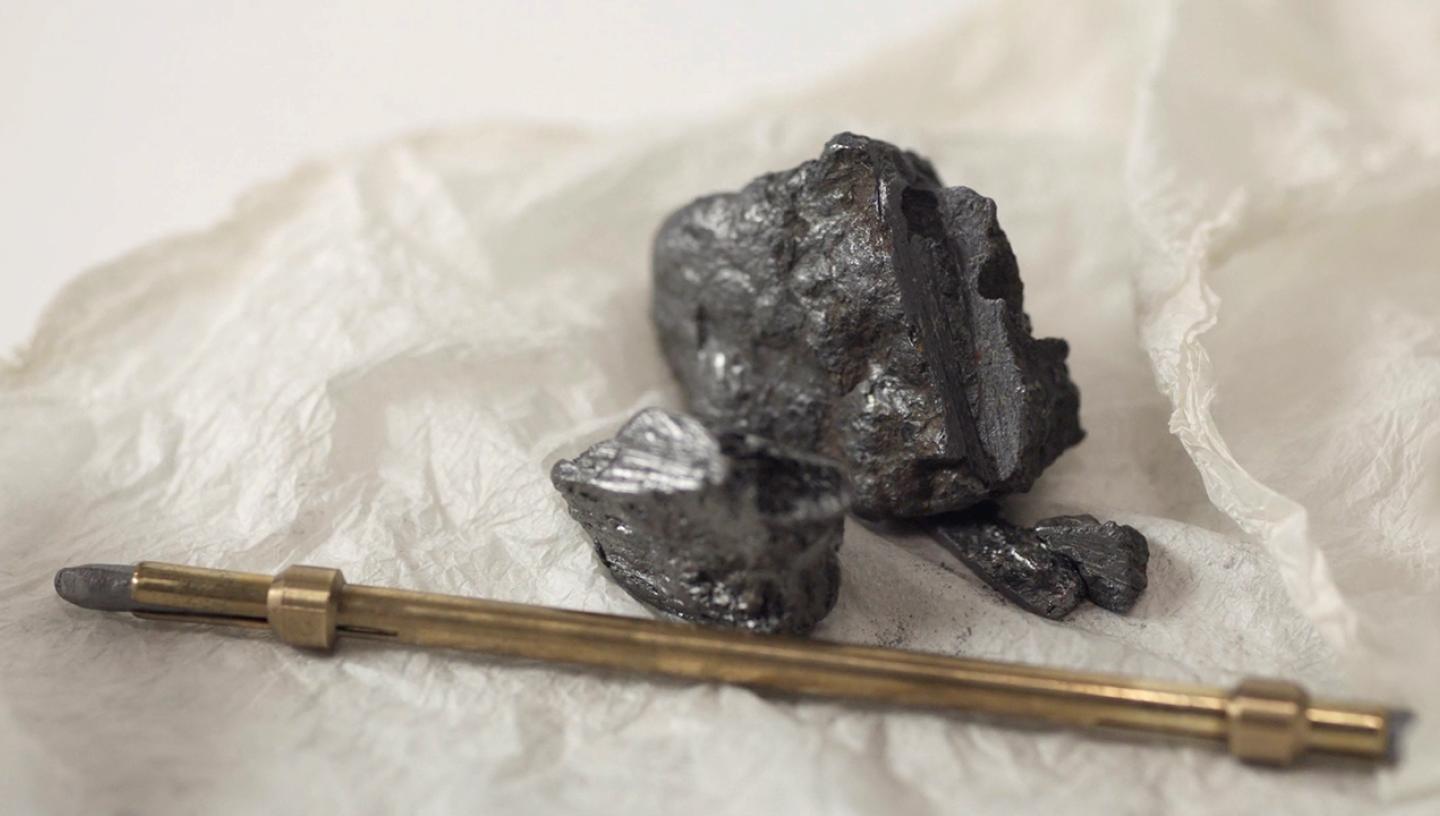 A lump of graphite and a metal lead holder on a piece of crumpled paper. The dark grey rock is shiny where it has been cut to be used for drawing, while the lead holder is a brass colour with a small nib of graphite sticking out of one end