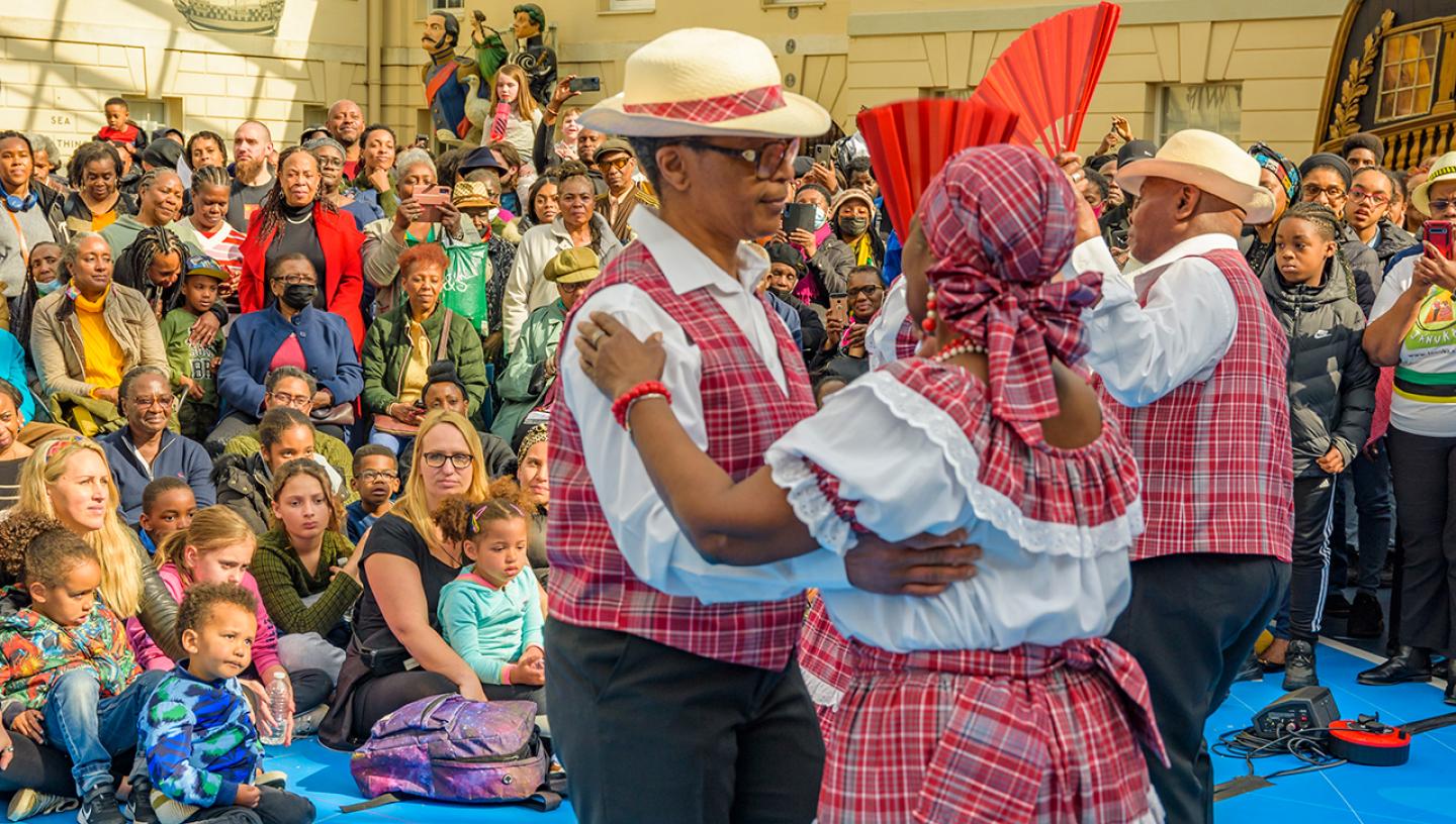 A quadrille dance performance, featuring a man and woman in bright red and white chequered cotton dancing arm in arm as a crowd look on