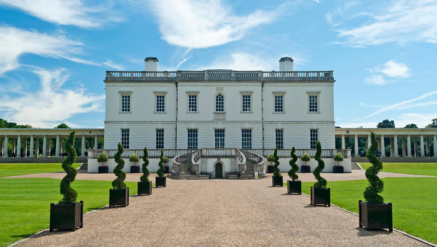 A view of the Queen's House in Greenwich from the outside. The square white building is almost symmetrical from the front, with a gravel pathway leading up to the main entrance