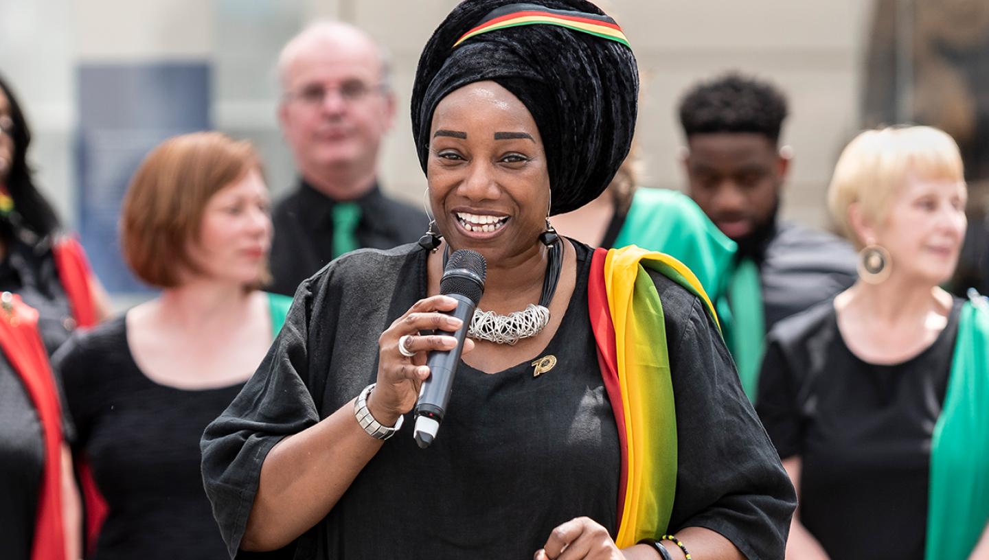 A female singer performs a solo as part of a choir performance. She is holding a microphone and is wearing a black dress with a multicoloured sash over her left arm, with a matching headscarf