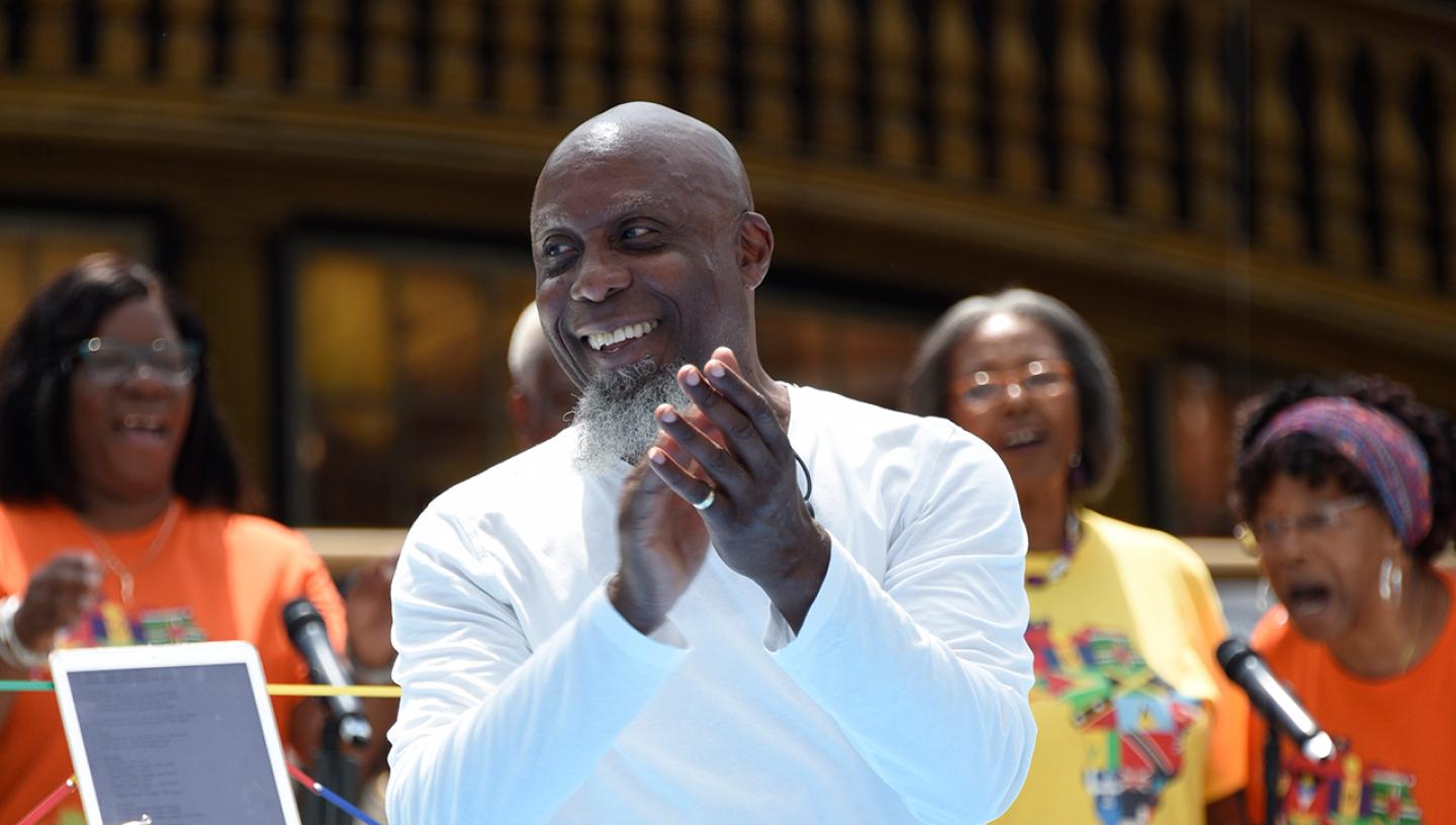 A man smiles and claps as he leads a choir during a performance at the National Maritime Museum. People in bright orange and yellow t-shirts can be seen singing in the background
